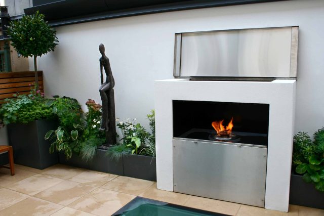 Small city garden design Knightsbridge with bespoke flambeaux fire burner and neat clipped planting, teak furniture parasol, clipped plants Hunza light fittings.