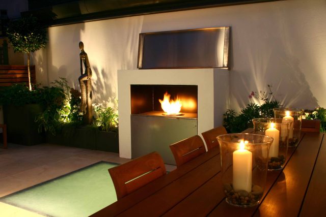 Small city garden design Knightsbridge with bespoke flambeaux fire burner and neat clipped planting, teak furniture parasol, clipped plants Hunza light fittings.