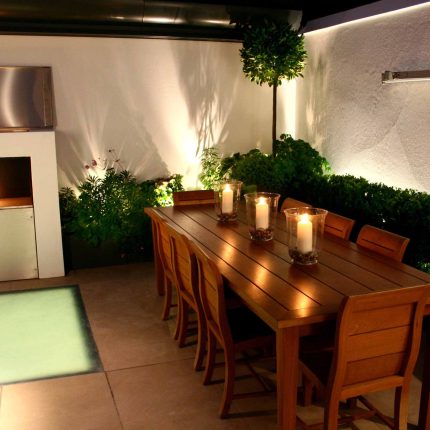 Small city garden design Knightsbridge with bespoke flambeaux fire burner and neat clipped planting, teak furniture parasol, clipped plants Hunza light fittings.
