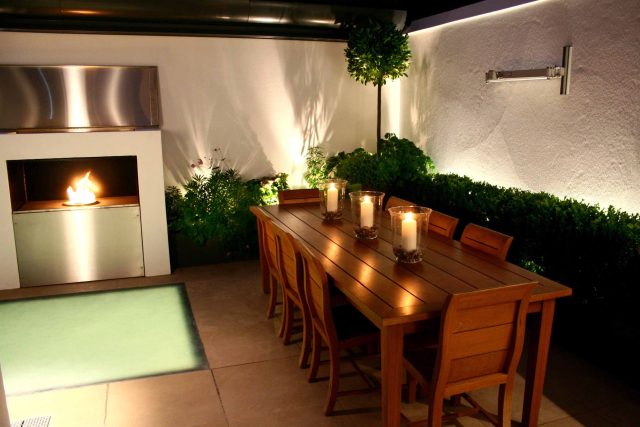 Small city garden design Knightsbridge with bespoke flambeaux fire burner and neat clipped planting, teak furniture parasol, clipped plants Hunza light fittings.