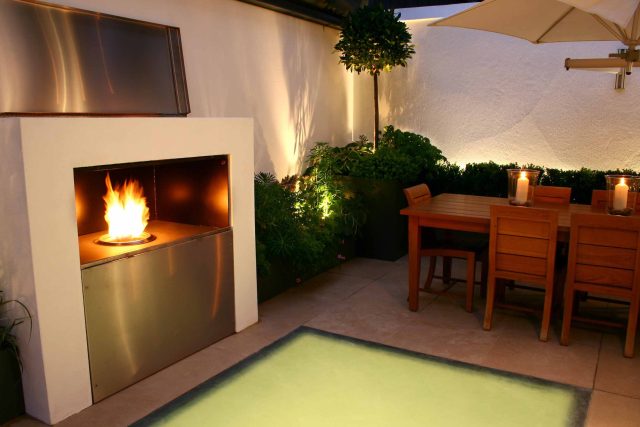 Small city garden design Knightsbridge with bespoke flambeaux fire burner and neat clipped planting, teak furniture parasol, clipped plants Hunza light fittings.