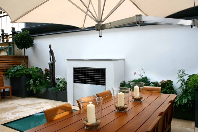 Small city garden design Knightsbridge with bespoke flambeaux fire burner and neat clipped planting, teak furniture parasol, clipped plants Hunza light fittings.