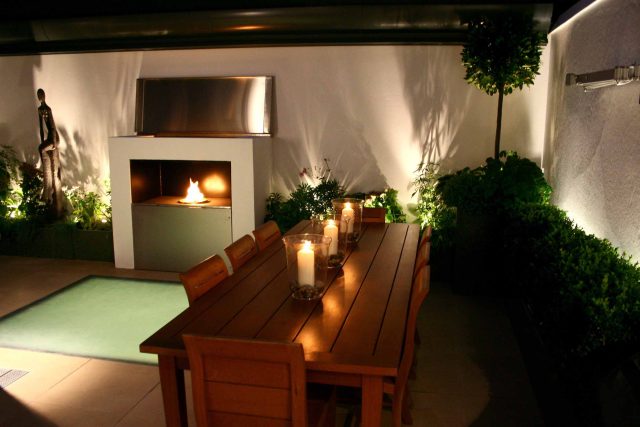 Small city garden design Knightsbridge with bespoke flambeaux fire burner and neat clipped planting, teak furniture parasol, clipped plants Hunza light fittings.