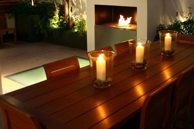 Small city garden design Knightsbridge with bespoke flambeaux fire burner and neat clipped planting, teak furniture parasol, clipped plants Hunza light fittings.