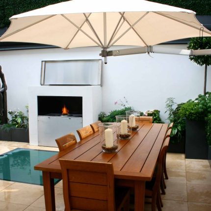 Small city garden design Knightsbridge with bespoke flambeaux fire burner and neat clipped planting, teak furniture parasol, clipped plants Hunza light fittings.