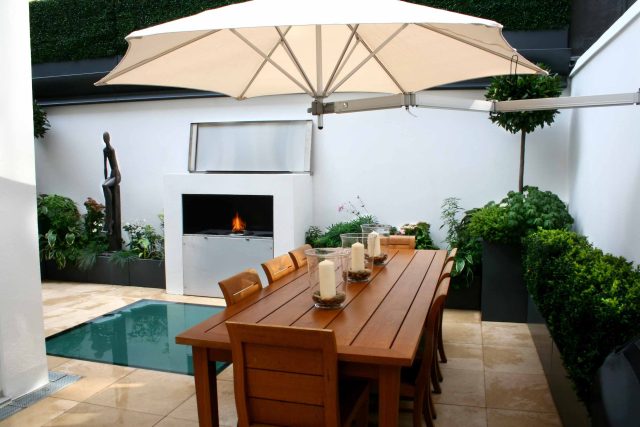 Small city garden design Knightsbridge with bespoke flambeaux fire burner and neat clipped planting, teak furniture parasol, clipped plants Hunza light fittings.