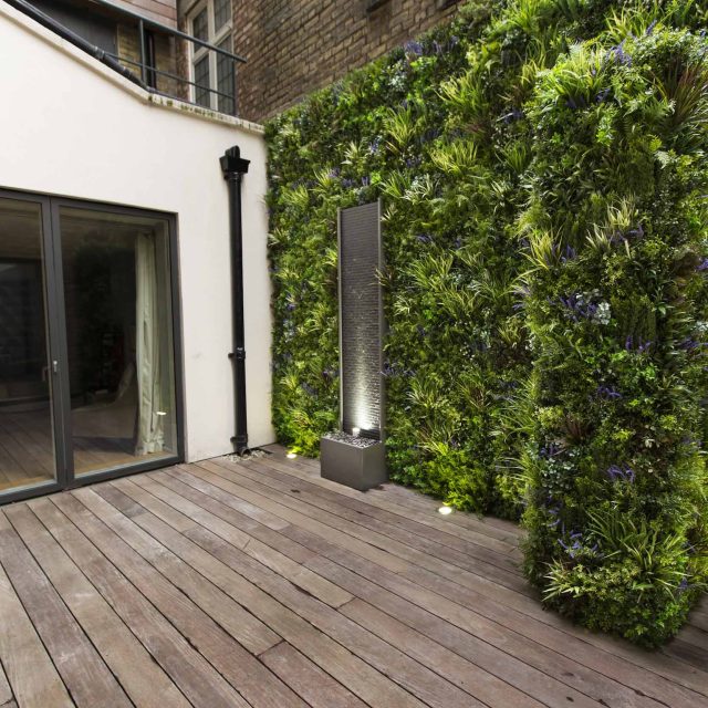 Small courtyard garden transformed with artificial green wall panels and a focal ribbed slate stone water feature, planting, natural light, small garden space.
