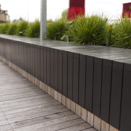 A black slat wood planter and bench on decking with ornamental grass.