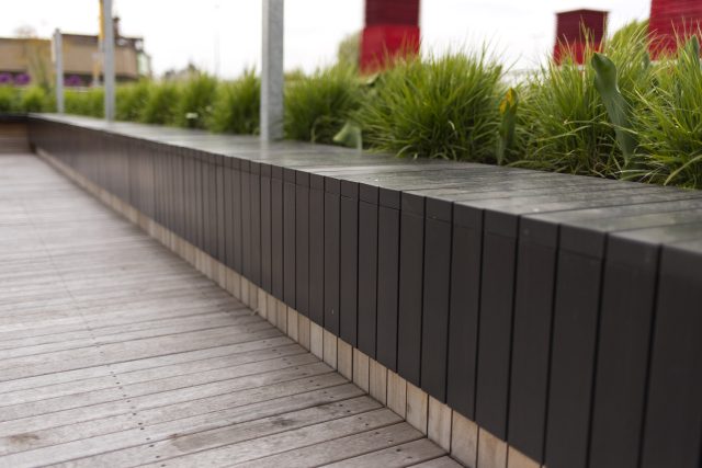 A black slat wood planter and bench on decking with ornamental grass.