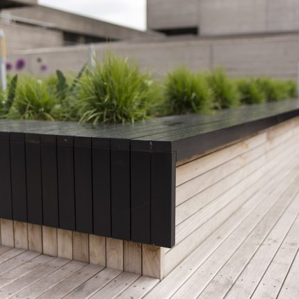 A modern black slat planter and bench with ornamental grass on decking.