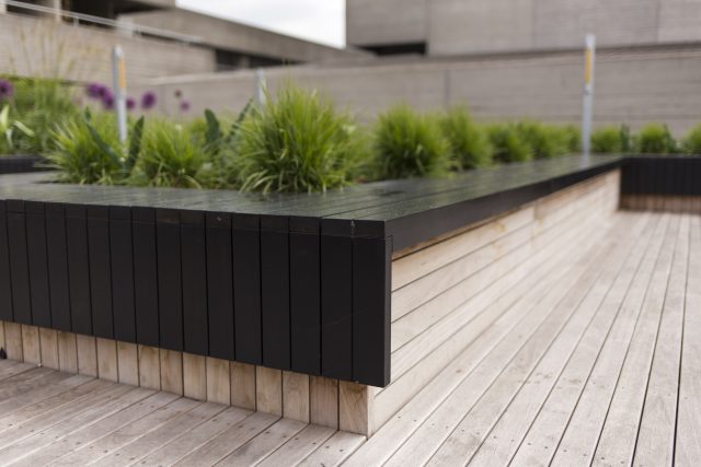 A modern black slat planter and bench with ornamental grass on decking.