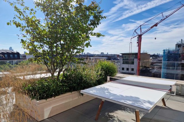 A ping pong table sits near a stone planter and grass in modern London.