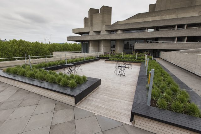 A wooden deck and grass planters on a brutalist London rooftop terrace.