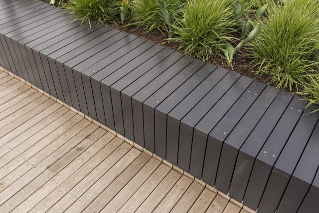 A slat bench, retaining wall, and wooden decking with ornamental grass.