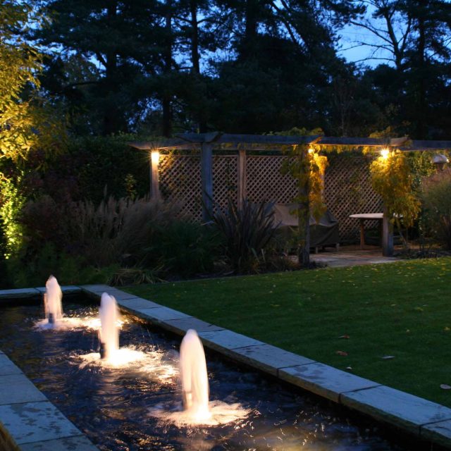Water Feature Garden design Surrey overgrown woodland space in Surrey with outdoor Hunza lighting designers mature planting, circular paving, cedar pergola.