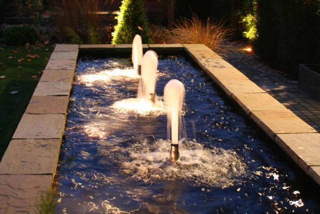 Water Feature Garden design Surrey overgrown woodland space in Surrey with outdoor Hunza lighting designers mature planting, circular paving, cedar pergola.