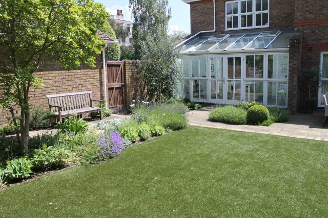 An artificial lawn, a brick conservatory, a patio, and topiary detail a formal yard.