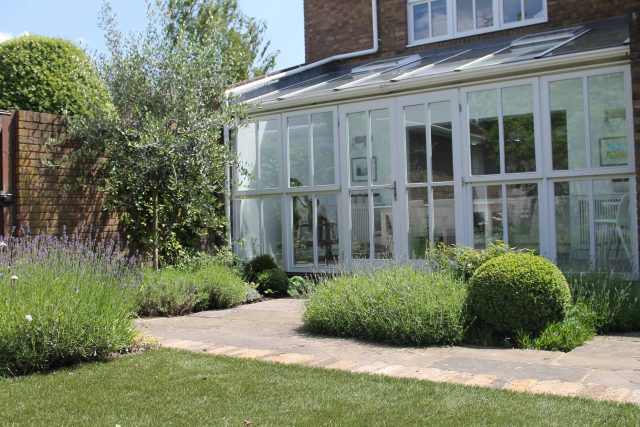 A conservatory opens to a stone patio with a lawn, boxwood topiary, and lavender.