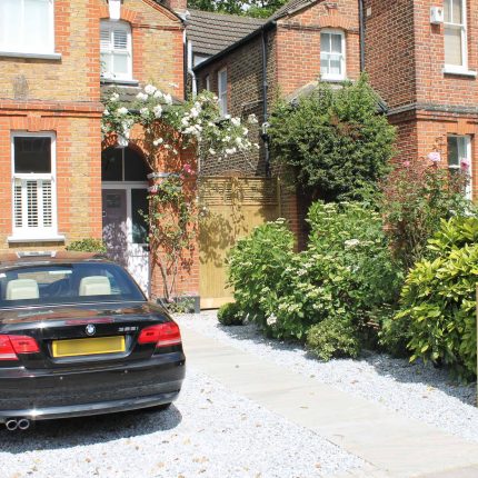 A gravel driveway and paved pathway lead past climbing roses and a slatted screen.