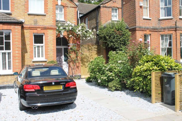 A gravel driveway and paved pathway lead past climbing roses and a slatted screen.