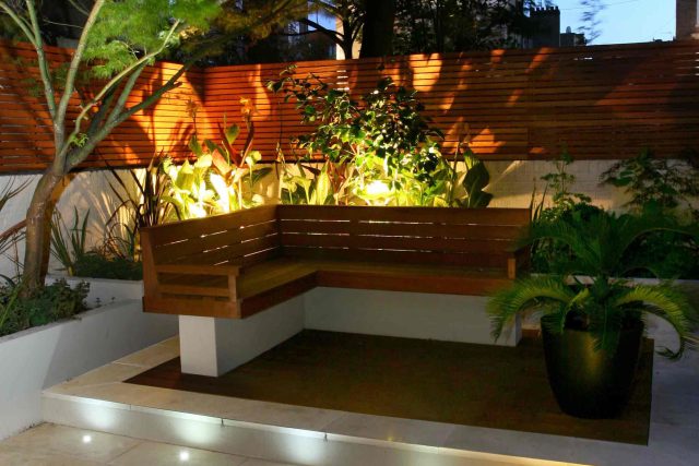 A hardwood bench and slatted fence illuminated by LED patio lighting on stone paving.
