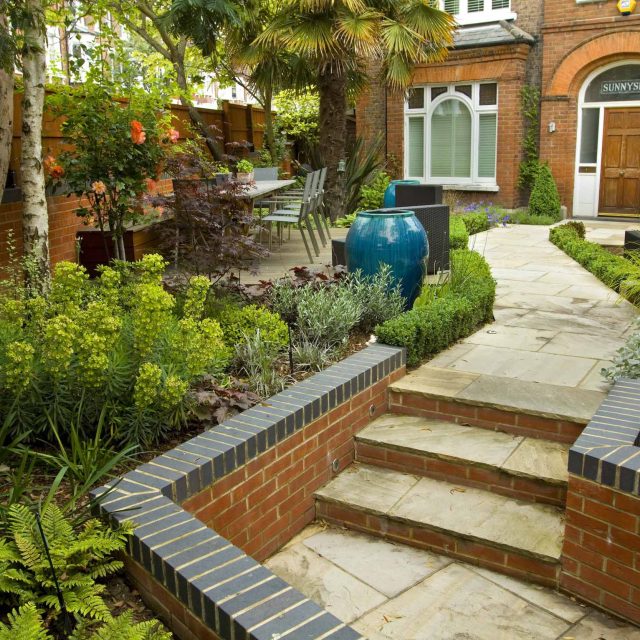Large paved garden terrace with sunken sandstone paved area, comfortable outdoor sofa by landscaping design company, raised timber decking, Garden Builders.