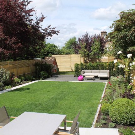 A modern stone patio is framed by a lawn, a slatted wood fence, and boxwood topiary.