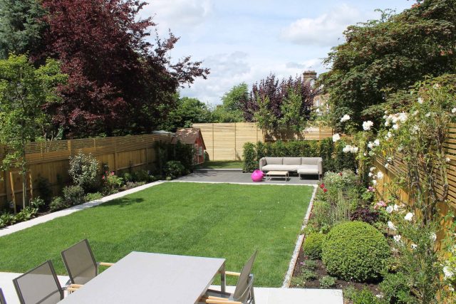 A modern stone patio is framed by a lawn, a slatted wood fence, and boxwood topiary.