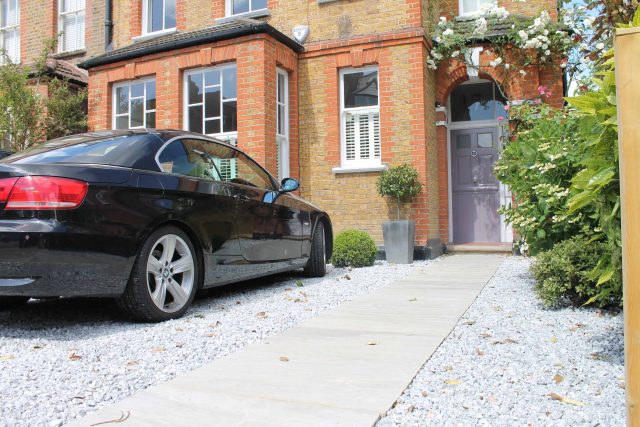 A stone slab pathway and gravel driveway sit in front of a brick house adorned with climbing roses.