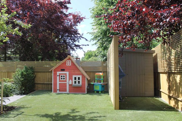 A wooden playhouse sits on artificial turf enclosed by a slatted timber privacy fence.
