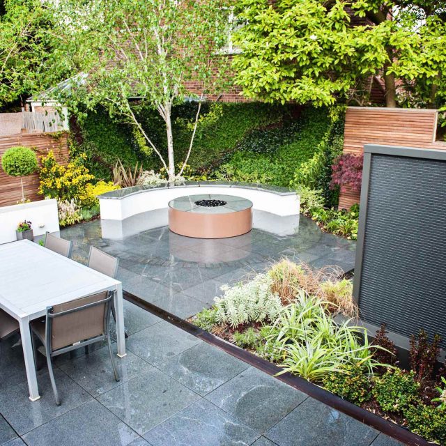 Suburban retreat design garden with circular stone fire table, artificial green wall surrounding natural planting, raised beds pillars artificial green walls.