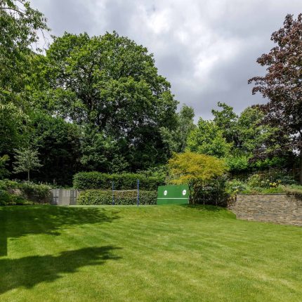 Large family garden design with outdoor kitchen, terrace, lighting, evergreens, lawn and stone-clad retaining wall designers specialist contractors in London.
