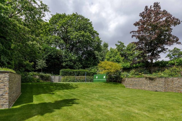 Large family garden design with outdoor kitchen, terrace, lighting, evergreens, lawn and stone-clad retaining wall designers specialist contractors in London.