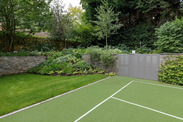 Large family garden design with outdoor kitchen, terrace, lighting, evergreens, lawn and stone-clad retaining wall designers specialist contractors in London.